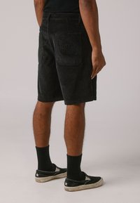Schwarze Cordshorts mit einer Gesäßtasche, die ein strukturiertes Emblem aufweist, in lässigem Schnitt und kombiniert mit schwarzen Socken und Slippern.