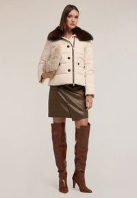 Giacca puff beige con colletto in pelliccia marrone, dettagli con bottoni, abbinata a una gonna in pelle marrone e stivali marroni al ginocchio con tacco.