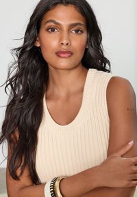 Krémový pletený top bez rukávov so širokým okrúhlym výstrihom, s rebrovanou textúrou a vrstvami náramkov v zlatej a čiernej farbe na zápästí.