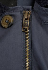 Primer plano de la cremallera de una chaqueta azul oscuro con un tirador de cremallera de latón que presenta una calavera y huesos cruzados, botón adyacente y detalles de la tela.