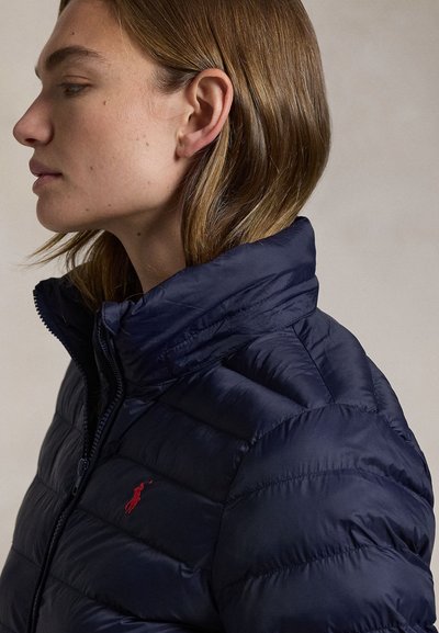 Heledate juustega naine kannab tumesinist tikitud jope kõrge kraega ja punase tikandiga rinnal, suunaga vasakule neutraalsel taustal.