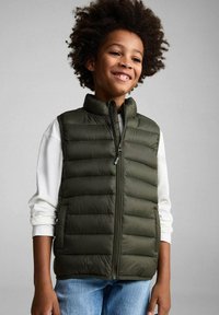 Zeleni puffer prsluk s visokim ovratnikom i zatvaranjem na zip, s horizontalnim quilted sekcijama. Nositi preko bijele majice s dugim rukavima.