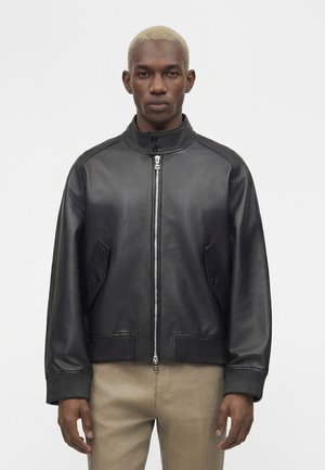 Homme aux cheveux blonds courts portant une veste en cuir noire zippée et un pantalon beige, debout devant un fond blanc uni.