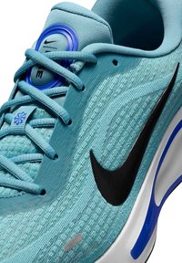 Sneakers Nike s lehkým modrým texturovaným svrškem, černým logem swoosh, modrými akcenty a elegantní bílou podrážkou se vzorovaným detailem.