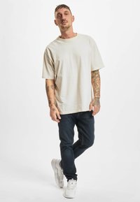 Světlé béžové oversized tričko z měkké látky, doplněné tmavě modrými džínami s úzkým střihem a bílými teniskami. Na pažích jsou viditelné tetování.