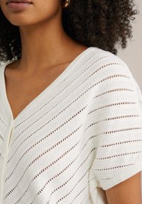 Kobieta ubrana w biały, krótki rękaw sweterka z poziomymi ażurowymi paskami i dekoltem w serek, pokazująca szyję, ramiona oraz kręcone włosy.