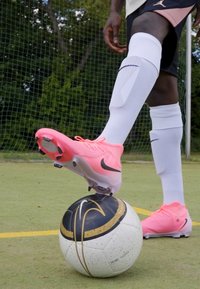 Ružičasta Nike nogometna kopačka odmara se na crno-bijelom nogometnom loptu s zlatnim akcentima, postavljena na zelenoj travi. Bijele čarape i crne kratke hlače su vidljive.