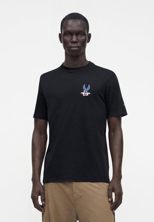 Bărbat purtând un tricou negru cu un mic logo colorat cu iepuraș de desene animate pe pieptul stâng și pantaloni bej, stând în fața unui fundal alb.