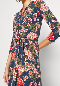 Kvinna i blå blommig klänning med röda, rosa blommor och gröna blad, med knappar framtill och knyt i midjan.