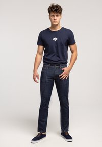 Młody mężczyzna z kręconymi włosami, ubrany w granatowy t-shirt z logo, ciemnoniebieskie dżinsy i granatowe sneakersy, stojący na jednolitym jasnym tle.