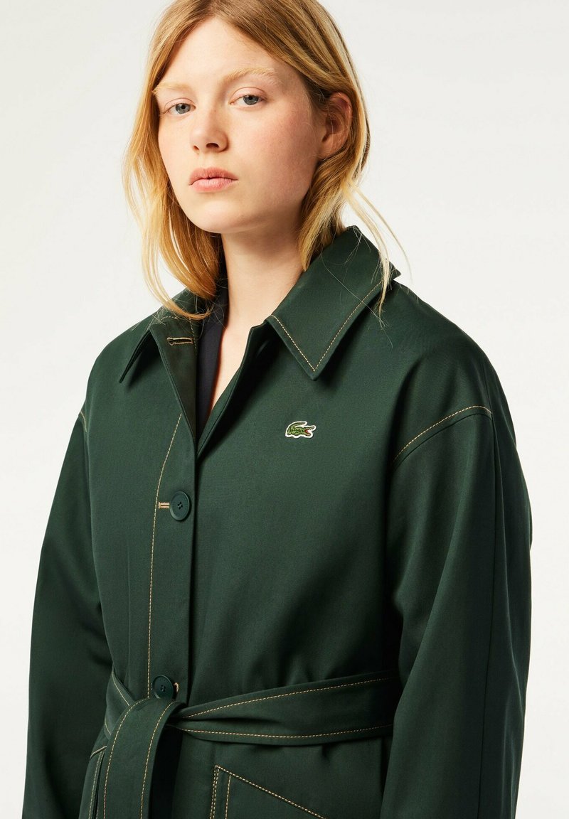 Manteau vert foncé au design ajusté, avec un grand col et des coutures visibles. Dispose d'une ceinture et d'un petit logo sur le côté gauche.