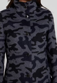 Wollen mantelblend met een grijs-zwart camouflagepatroon, hoge kraag en vijf glanzende zwarte knopen aan de voorkant.