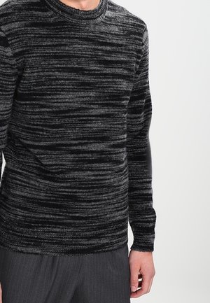 Mand iført en langærmet sort og gråstribet strikket sweater med mørkegrå bukser med smalle striber foran en ensfarvet baggrund.