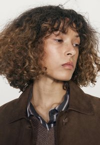 Bruine suède jas met drukknoopsluiting, gedragen over een gestreept shirt. Het model heeft krullend haar met een natuurlijke textuur.