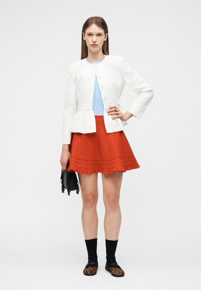 Γυναίκα που φοράει λευκό σακάκι με κουμπιά, μπλε ριγέ τοπ, πορτοκαλί πλισέ φούστα, μαύρες κάλτσες, παπούτσια με λεοπάρ σχέδιο, κρατώντας μια μαύρη τσάντα με φούντες.