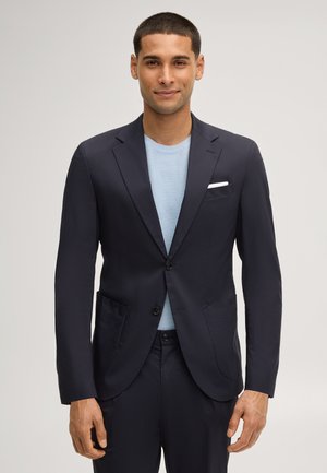 Man in een marineblauw pak met een lichtblauw gestructureerd overhemd, staand en voorwaarts kijkend tegen een effen achtergrond.