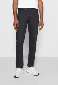 En person står i en neutral miljö, klädd i en vit t-shirt och mörka jeans, tillsammans med vita sneakers. Händerna avslappnade vid sidorna.