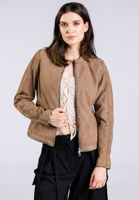 Giacca in pelle marrone con scollatura rotonda, chiusura con cerniera, tasche laterali e dettagli sulle cuciture, indossata sopra un leggero top all'uncinetto.