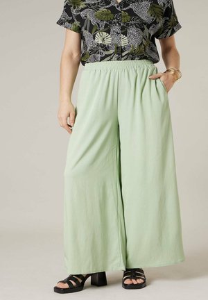 Femme portant un pantalon large vert clair, des sandales à talons carrés noires à brides, et un chemisier noir à fleurs à manches courtes, une main dans la poche.