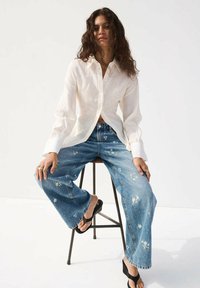Mujer con cabello rizado y castaño sentada en un taburete, lleva una camisa blanca de manga larga, vaqueros de pierna ancha con bordado floral y sandalias negras.