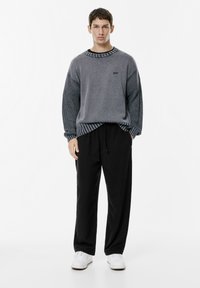 Szary sweter z fakturowanymi rękawami, prążkowanym dekoltem i czarnymi spodniami; model stoi w białych trampkach na jednolitym tle.