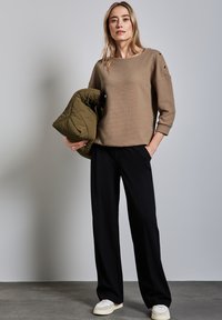 Beiger, strukturierter Pullover mit Dreiviertelärmeln, kombiniert mit schwarzen, weiten Hosen und einer olivgrünen Steppjacke, die im linken Arm gehalten wird.