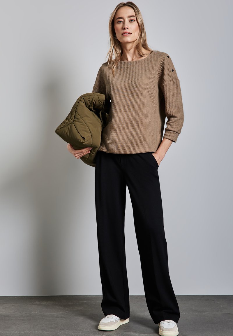 Beiger, strukturierter Pullover mit Dreiviertelärmeln, kombiniert mit schwarzen, weiten Hosen und einer olivgrünen Steppjacke, die im linken Arm gehalten wird.