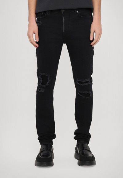 Jean en denim noir avec une coupe slim, présentant des patchs déchirés aux genoux, un design classique à cinq poches et une fermeture à bouton à la taille.