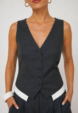 Donna che indossa un gilet senza maniche grigio scuro con bottoni e scollo a V profondo, orecchini a cerchio dorati, bracciale bianco e pantaloni grigio scuro coordinati.