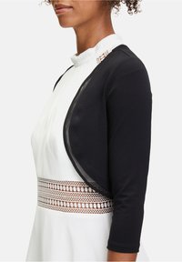 Musta bolero-takki, jossa on läpikuultava reunus, käytetty valkoisen mekko päällä, jossa on poimutuksia ja koristeellinen pitsivyötärö. Lyhennetyt hihat, sileä tekstiili.