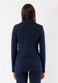 Blazer aderente blu navy con rever a punte, maniche lunghe e una texture liscia, caratterizzato da cuciture che enfatizzano la silhouette.