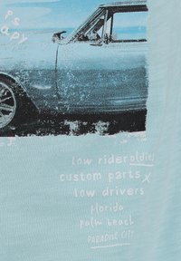 Camiseta de color azul claro con un gráfico de un coche antiguo azul, el texto incluye "Modelos lowrider," "piezas personalizadas," y "Ciudad del Paraíso."