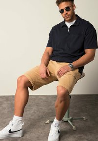 Polo shirt blu navy con zip e maniche corte, pantaloni corti khaki, sneakers bianche Nike e occhiali da sole neri. Seduto su uno sgabello.