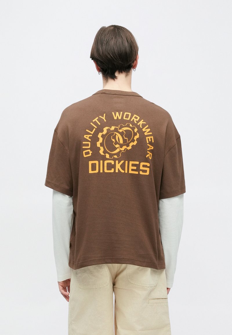 Personne aux cheveux courts portant un t-shirt de travail Dickies marron par-dessus une chemise blanche à manches longues et un pantalon beige, tournée de dos à la caméra.