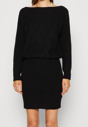 Femme portant une robe noire en maille texturée à manches longues avec un collier de perles, vue des épaules jusqu'au milieu des cuisses.