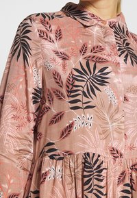 Robe en tissu léger et rose avec un col boutonné, ornée de motifs de feuilles noirs et corail. Taille froncée et manches fluides.