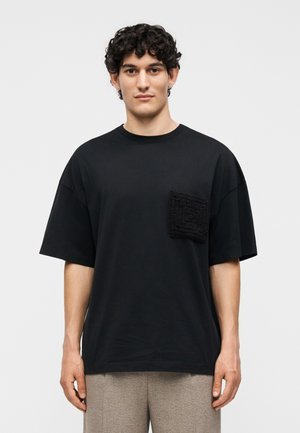 Νεαρός άνδρας με σγουρά σκούρα μαλλιά που φοράει φαρδιά μαύρη μπλούζα με υφασμένη μπροστινή τσέπη και ανοιχτόχρωμο παντελόνι, στέκεται μπροστά σε απλό φόντο.