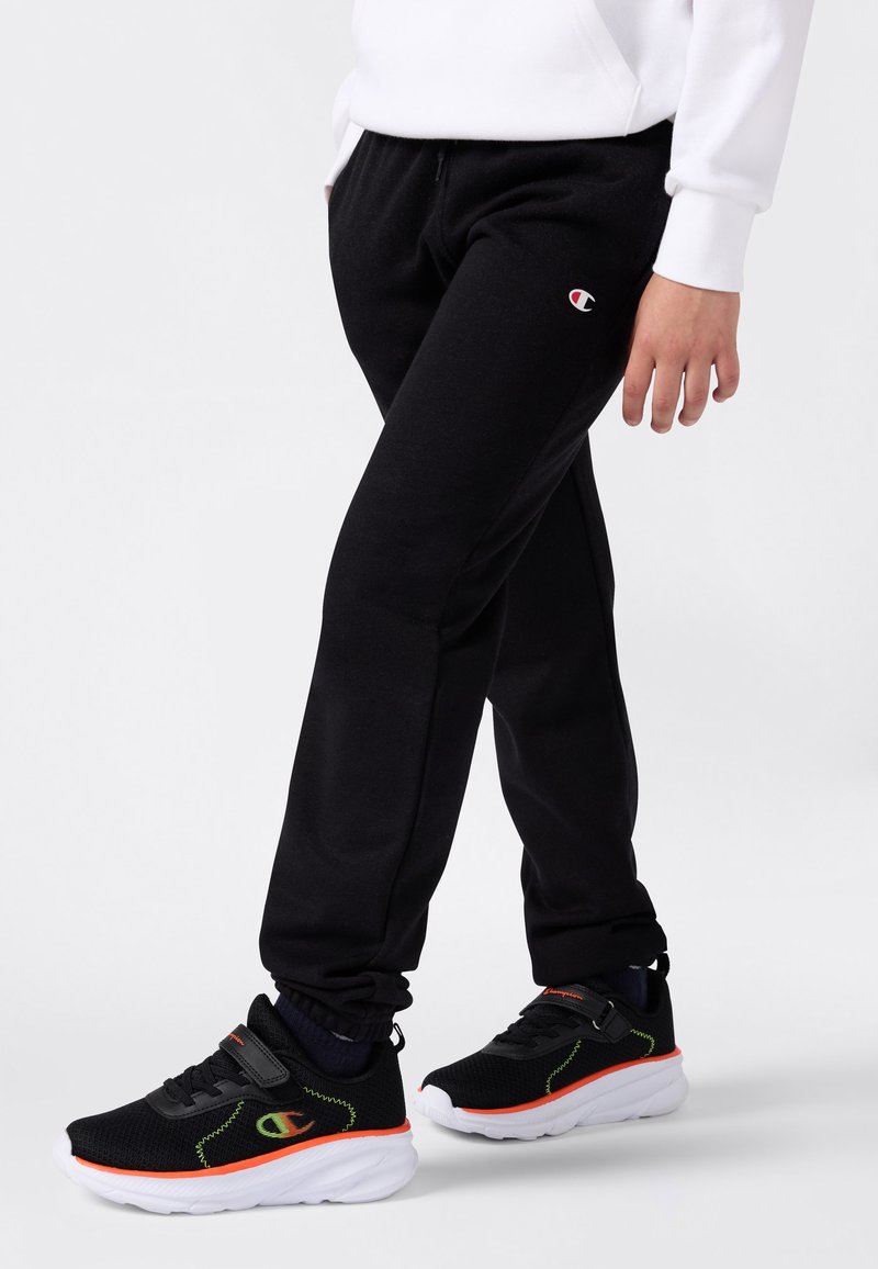 Person, der schwarze Champion-Sweatpants und schwarze Sneakers mit orangefarbenen und grünen Details trägt, steht vor einem schlichten weißen Hintergrund.
