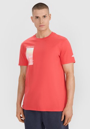 Mężczyzna z krótkimi włosami, ubrany w koralowo-czerwony T-shirt z małą grafiką i białym tekstem w górnym lewym rogu oraz ciemnoniebieskie spodnie.