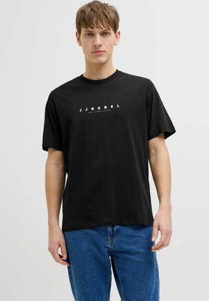 Joven con camiseta negra con el texto "JJREBEL" y vaqueros azules, de pie frente a un fondo claro y sencillo.