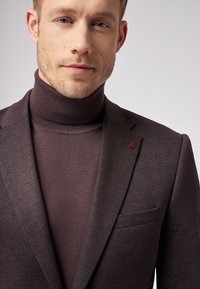 Ciemnobrązowa marynarka uszyta z teksturowanej tkaniny, z klapami typu notch i małym czerwonym logo na klapie, noszona na dopasowanym golfie.