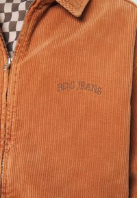 Kurtka sztruksowa w spalonym pomarańczowym kolorze z pionowymi żebrowaniami, z zamkiem błyskawicznym i haftem "BDG JEANS" w ciemnym niciu nad suwakiem.