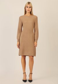 Strickkleid in Hellbraun, mit einem strukturierten Zopfmuster im Oberteil, langen Ärmeln und einem ausgestellten Saum, kombiniert mit schwarzen High Heels.