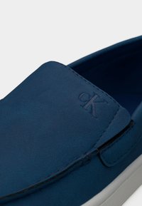 Chaussure bleu daim à enfiler avec une texture lisse, un bout en forme d'amande, une semelle blanche et un détail de logo en relief sur le côté.