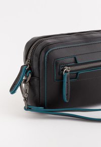 Bolso de muñeca de cuero negro con ribete azul, cremalleras plateadas, bolsillo frontal con cremallera y correa de muñeca desmontable sobre un fondo blanco.