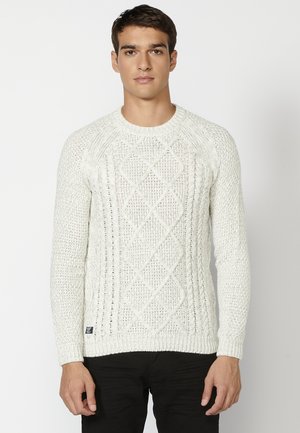 Suéter blanco de punto con un patrón de diamantes, puños acanalados y cuello redondo. El hilo texturizado y grueso le da un aspecto acogedor.
