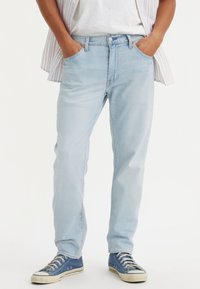 Lichtblauwe spijkerbroek met een rechte pijp, vijf zakken en metalen knoopsluiting. Draagt bij blauwe sneakers en een gestreept shirt.