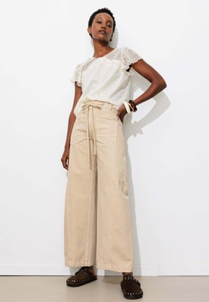 Femme portant un chemisier blanc transparent à manches courtes, un pantalon beige large avec une ceinture à nouer, des chaussures sombres sans lacets et des bracelets épais à un poignet.