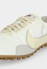 Λευκό και μπεζ αθλητικό παπούτσι Nike με εκρού κορδόνια, ραμμένο λογότυπο swoosh και σόλα από καουτσούκ χρώματος καραμέλας, ορατή σε φωτεινό φόντο.
