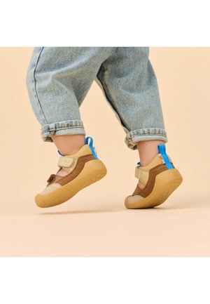 Kind draagt beige en bruine schoenen met blauwe trekbandjes en opgerolde spijkerbroek, halverwege een stap op een beige achtergrond.